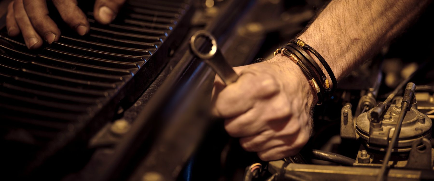 Mãos de mecânico apertando uma peça do motor com chave de boca, sob luz amarelada